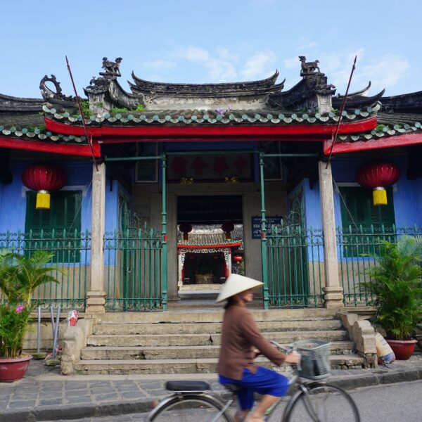 Eine Vietnamesin mit konischem Hut fährt auf einem Fahrrad an einem chinesischen Tempel vorbei.