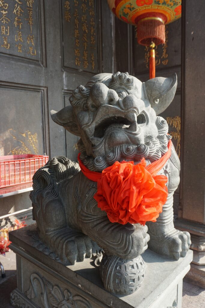 Steinfigur eines Löwen, der den Eingang zu einem chinesischen Tempel bewacht. Er trägt eine große rote Schleife um den Hals.